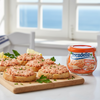 Bocadelia jar with salmon pate on bread slices on a wooden board by a window.